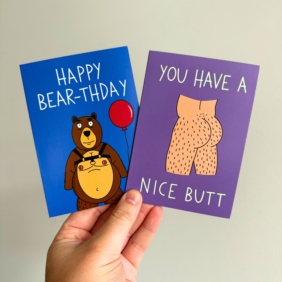 Two greeting cards held in a hand, one with a cartoon bear holding a balloon reading "HAPPY BEAR-THDAY" and the other with a humorous text 'YOU HAVE A NICE BUTT' accompanied by a bear's rear end illustration.